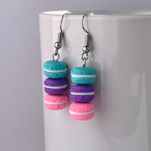 May include: Dangle earrings featuring three stacked, colourful macarons. Each macaron is a different colour: teal, purple, and pink, with white filling. The earrings have silver-coloured hooks and springs.