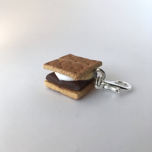 May include: A miniature s'more keychain charm featuring a graham cracker, chocolate, and marshmallow. The charm is attached to a silver metal lobster clasp.