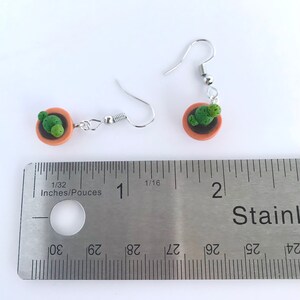 Puede incluir: Un par de pendientes de plata con un peque&ntilde;o cactus en maceta. Los pendientes miden aproximadamente 2,5 cm de largo. El cactus es verde y la maceta es marr&oacute;n.