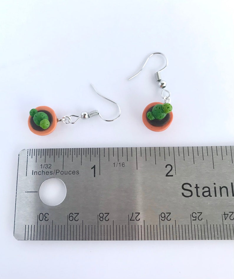 Puede incluir: Un par de pendientes de plata con un peque&ntilde;o cactus en maceta. Los pendientes miden aproximadamente 2,5 cm de largo. El cactus es verde y la maceta es marr&oacute;n.