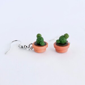 Puede incluir: Un par de pendientes colgantes con cactus en maceta en miniatura. Los pendientes est&aacute;n hechos de arcilla polim&eacute;rica y tienen ganchos plateados.