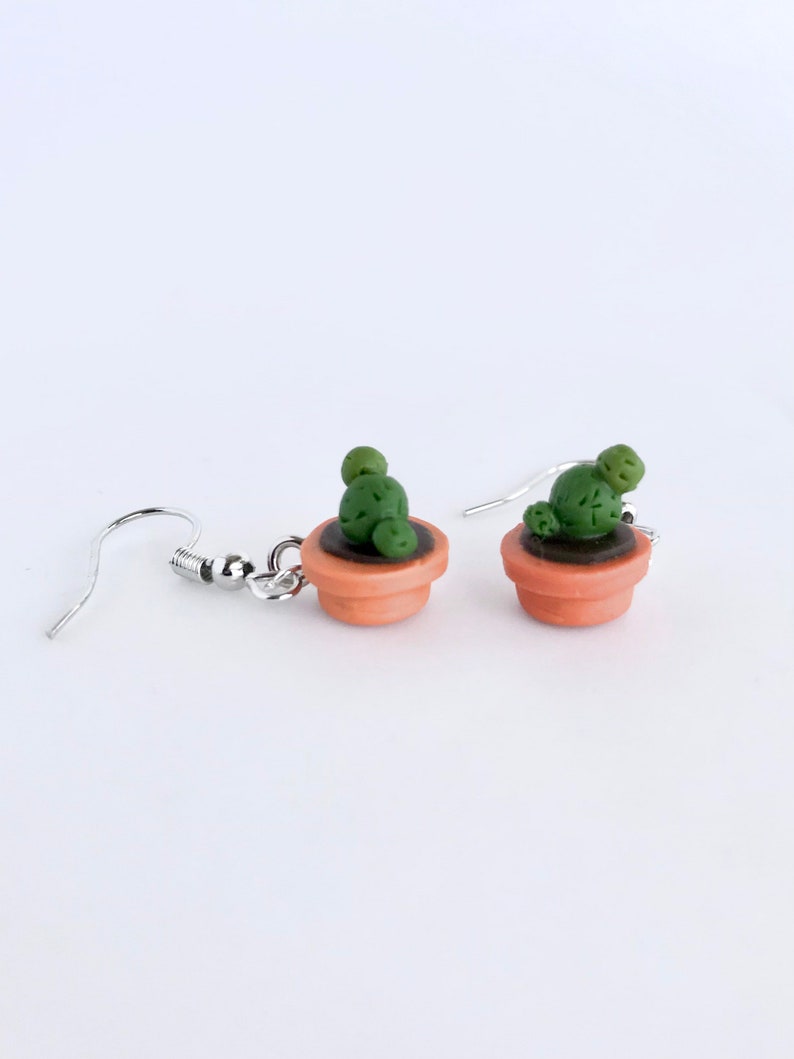 Puede incluir: Un par de pendientes colgantes con cactus en maceta en miniatura. Los pendientes est&aacute;n hechos de arcilla polim&eacute;rica y tienen ganchos plateados.