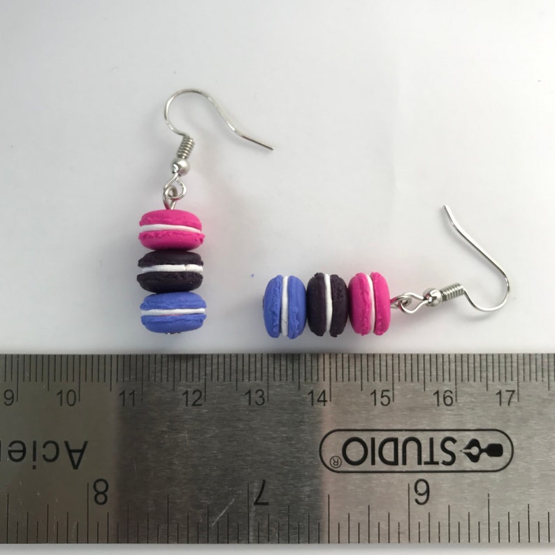 May include: A pair of dangle earrings featuring three stacked macarons. The macarons are pink, black, and blue with white frosting. The earrings are approximately 1.5 inches long.