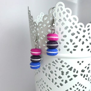 May include: A pair of dangle earrings featuring three stacked macarons in pink, purple and blue with white frosting.