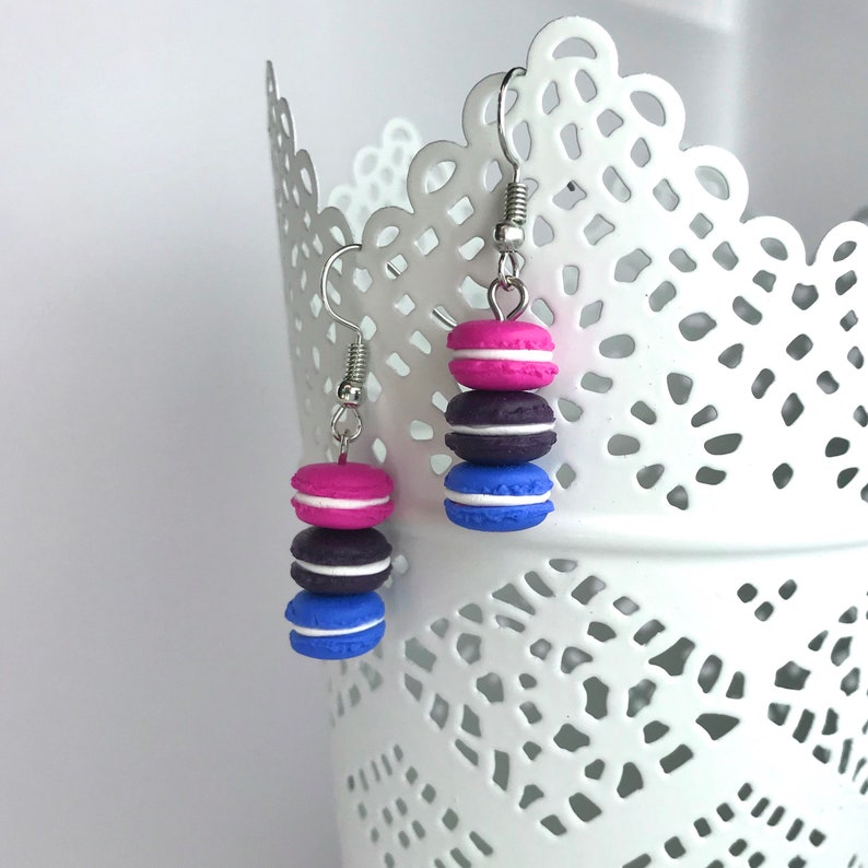 May include: A pair of dangle earrings featuring three stacked macarons in pink, purple, and blue with white frosting.