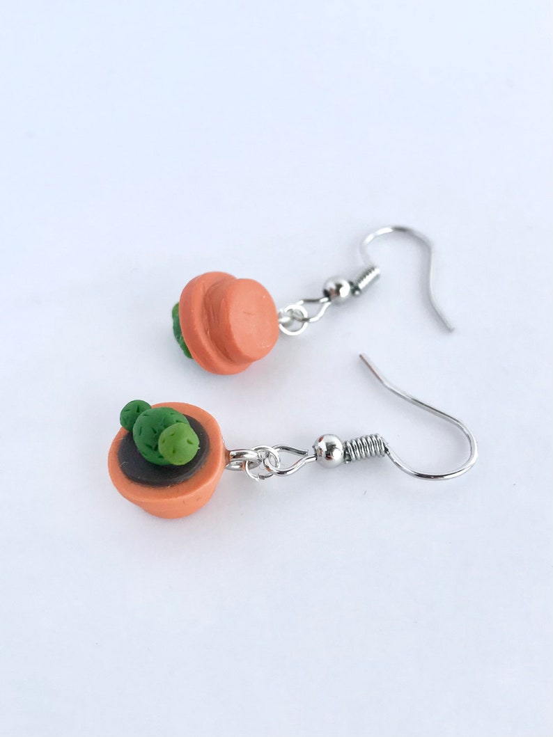 Puede incluir: Un par de pendientes de aro de plata con un peque&ntilde;o encanto de maceta de terracota con una planta de cactus verde.