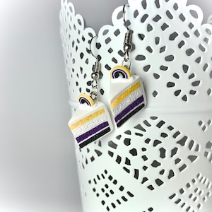 Peut inclure: Une paire de boucles d'oreilles pendantes représentant une part de gâteau avec des couches jaunes, blanches, violettes et noires. Le gâteau est surmonté d'un tourbillon jaune et violet.