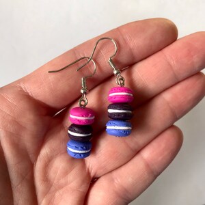 May include: A pair of dangle earrings featuring three stacked macarons in pink, black, and blue colors. The macarons have white frosting and are attached to silver hooks.