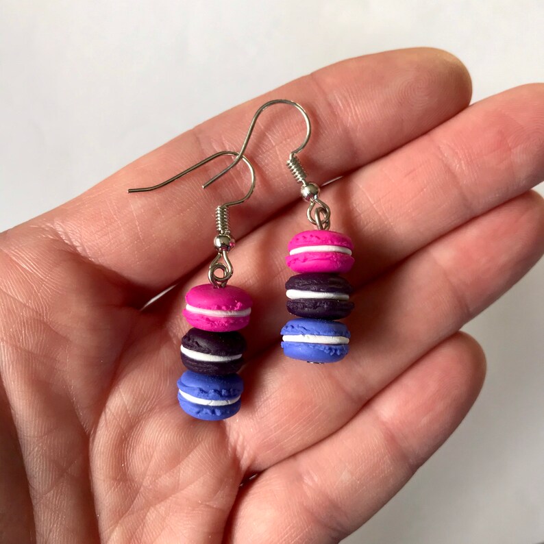 May include: A pair of dangle earrings featuring three stacked macarons in pink, black, and blue colors. The macarons have white frosting and are attached to silver hooks.