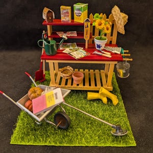 Puede incluir: Escena de jardín en miniatura con un banco de macetas rojo y de madera natural, una carretilla y herramientas. Incluye una regadera, girasoles y una pequeña calabaza. La escena está sobre un trozo de hierba verde.