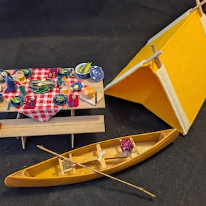 Op de afbeelding: Miniatuur kampeerscène met een gele tent, een houten kano met roeispanen en een picknicktafel gedekt voor een maaltijd. De tafel heeft een rood-wit geblokt tafelkleed en is gedekt met borden, kommen en eten. Er is ook een rode lantaarn aanwezig.