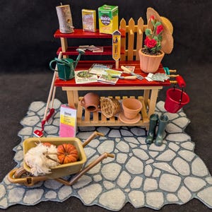 Puede incluir: Escena de jardín en miniatura con un banco de macetas rojo y de madera, completo con herramientas, plantas y una carretilla. La escena incluye una regadera, una rosa en maceta y una pequeña carretilla llena de calabazas y heno. Una escena de jardín en miniatura.