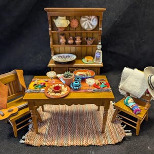 Może przedstawiać: Miniaturowa scena jadalni ze stołem drewnianym nakrytym do posiłku. Zawiera małą komodę, krzesła, dywan i różne produkty spożywcze. Gitara i kamizelka znajdują się na krześle. Sombrero i lalka znajdują się na innym krześle.