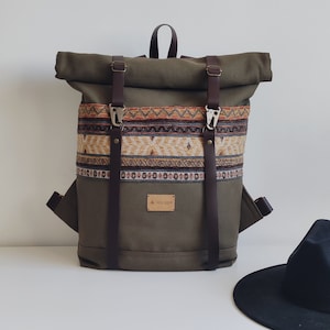 May include: A green canvas backpack with brown leather straps and a roll-top closure. The backpack has a woven patterned band with red, orange, and brown accents. The brand name "A.HILVAN" is printed on a brown leather tag.