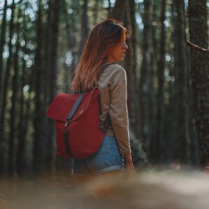 May include: A woman wearing a beige sweater and denim shorts is standing in a forest. She is carrying a red backpack with black straps.