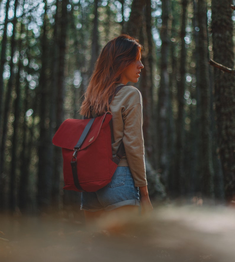 May include: A woman wearing a beige sweater and denim shorts is standing in a forest. She is carrying a red backpack with black straps.
