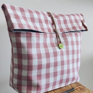 Könnte beinhalten: Eine rosa-weiß karierte Stofftasche mit gerolltem Oberteil, gesichert durch einen Kordelzug und einen grünen Knopf. Die Tasche steht auf einem Holzschemel, mit einem Häkelhaken und Knöpfen.