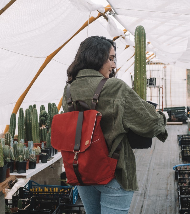 May include: A woman wearing a green corduroy jacket carries a red canvas backpack with brown leather straps and buckles. The backpack is worn over her shoulder and has a flap closure. The woman is standing in a greenhouse with various cacti plants in the background.