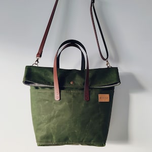 Könnte beinhalten: Olivgrüne Tragetasche mit braunen Ledergriffen und einem abnehmbaren Schultergurt. Die Tasche hat einen Überschlag und ein kleines Lederetikett mit Text. Die Tasche hängt vor einem weißen Hintergrund.