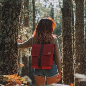 May include: A woman with long brown hair wearing a red canvas backpack with black straps and blue denim shorts stands in a forest.