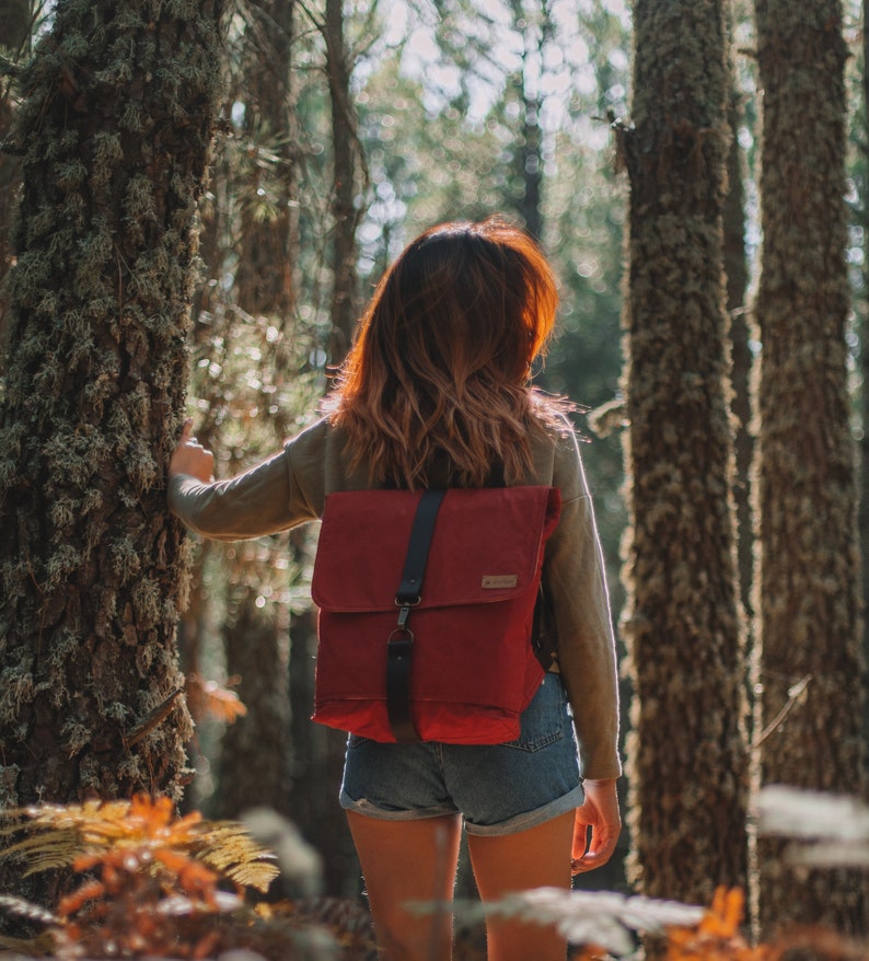 May include: A woman with long brown hair wearing a red canvas backpack with black straps and blue denim shorts stands in a forest.