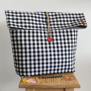 Op de afbeelding: Een zwart-wit geruite stoffen draagtas met een opgerolde bovenkant, vastgezet met een bruin koord en een rode knoop. De tas staat op een kleine houten kruk. Een paar knopen en een haaknaald liggen op de kruk.