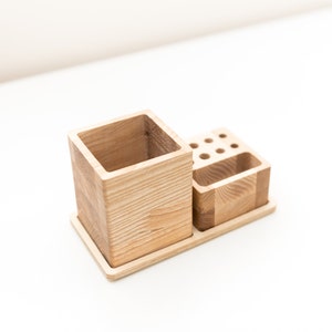 May include: A wooden desk organizer with a square pen holder, a small compartment, and a section with multiple holes. The organizer is made of light-colored wood and sits on a rectangular base.