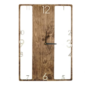 May include: A wooden wall clock with a brown wood grain finish. The clock face is surrounded by a white frame with cut-out numbers. The numbers are in a white font and are arranged in a circle around the clock face. The clock hands are black.