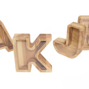 Pu&ograve; includere: Quattro salvadanai di legno a forma delle lettere A, J, K e B. I salvadanai sono realizzati in legno marrone chiaro e hanno un inserto in plastica trasparente nella parte anteriore.