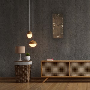 May include: A wooden clock with white numbers and a black hand, hanging on a gray wall. A wooden side table with a wicker basket and a lamp sits in front of the clock. Two pendant lights hang above the table.