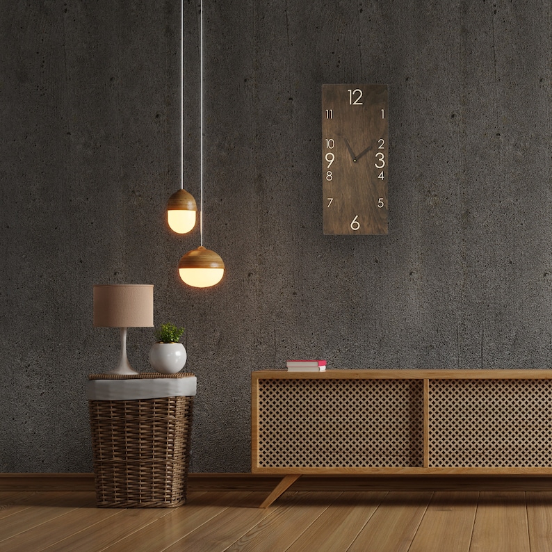 May include: A wooden clock with white numbers and a black hand, hanging on a gray wall. A wooden side table with a wicker basket and a lamp sits in front of the clock. Two pendant lights hang above the table.