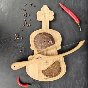Könnte beinhalten: Ein hölzernes Brotbrett in Geigenform mit einer Brotscheibe und einem Brotschneider. Eine rote Chilischote und Kaffeebohnen befinden sich ebenfalls auf der dunklen Oberfläche. Auf dem Brett steht das Wort "promi".