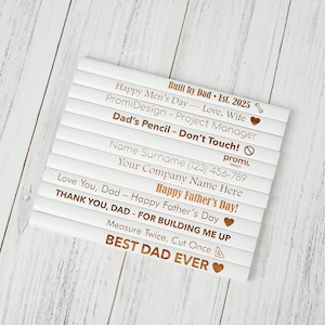 May include: A set of white pencils with gold lettering. The pencils have various phrases such as "Happy Father's Day!" and "Best Dad Ever." The pencils are arranged on a light wood surface.