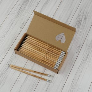 Pode incluir: Uma caixa de cartão com lápis personalizados, sobre uma superfície de madeira clara. Os lápis são de madeira natural com bandas e borrachas prateadas. A caixa tem um recorte em forma de coração.
