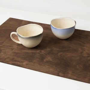 May include: Two ceramic mugs on a brown wooden placemat. One mug is white with a blue glaze, the other is white with a gray glaze.