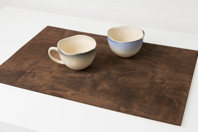 May include: Two ceramic mugs on a brown wooden placemat. One mug is white with a blue glaze, the other is white with a gray glaze.