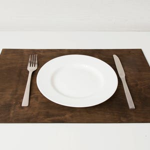 May include: A white ceramic dinner plate with a silver fork and knife on a dark brown wooden placemat.