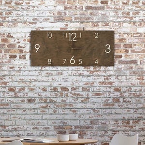 K&ouml;nnte beinhalten: Eine braune Holzuhr mit wei&szlig;en Zahlen an einer wei&szlig;en Backsteinwand. Die Uhr h&auml;ngt an der Wand und die Zeiger zeigen die Zeit an. Vor der Wand steht ein Holztisch mit zwei wei&szlig;en St&uuml;hlen.