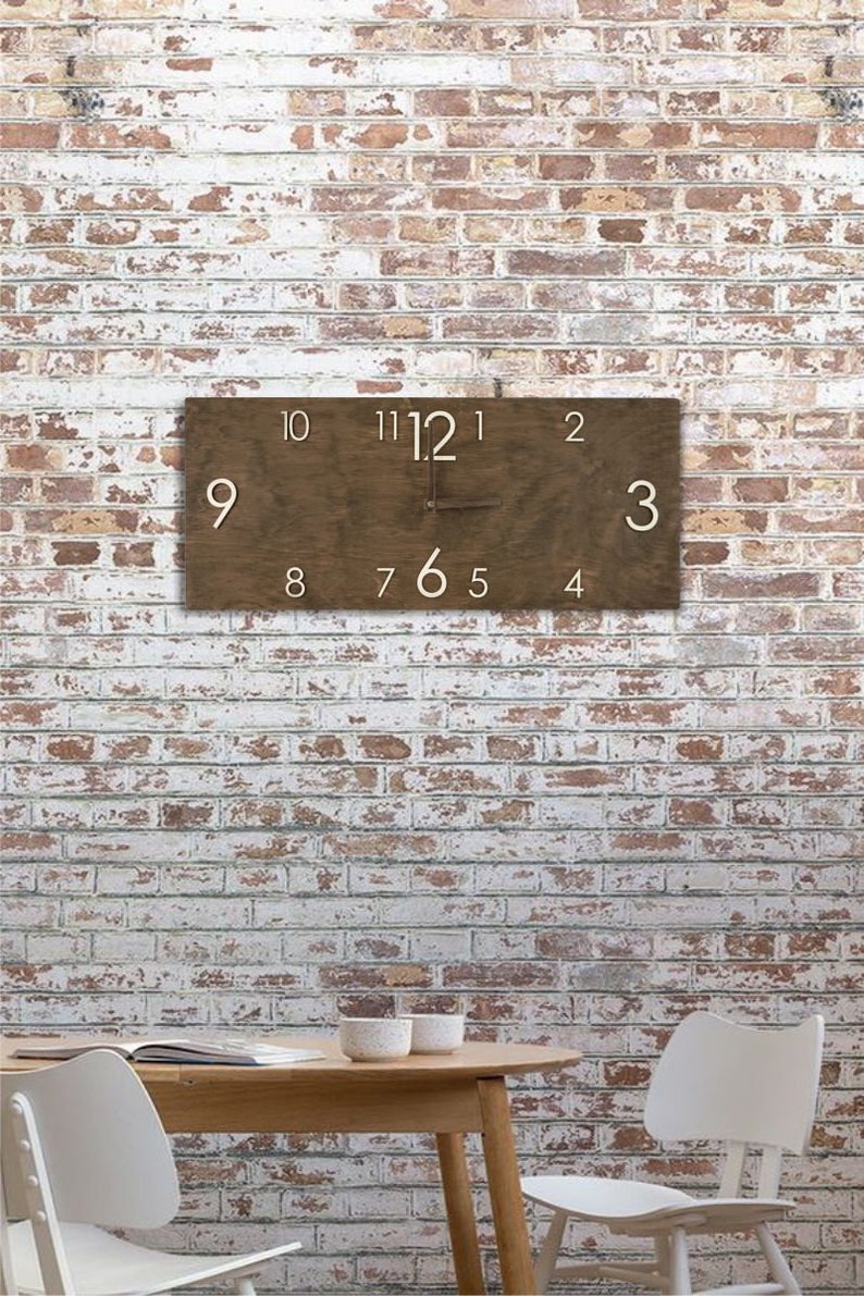K&ouml;nnte beinhalten: Eine braune Holzuhr mit wei&szlig;en Zahlen an einer wei&szlig;en Backsteinwand. Die Uhr h&auml;ngt an der Wand und die Zeiger zeigen die Zeit an. Vor der Wand steht ein Holztisch mit zwei wei&szlig;en St&uuml;hlen.
