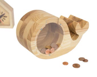 Puede incluir: Huchita de madera en forma de ballena con una ranura para monedas de plástico transparente. La huchita es de color marrón claro y tiene algunas monedas dentro.