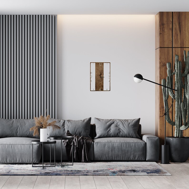 May include: A gray sofa with two throw pillows and a black throw blanket sits in a living room with a wooden wall and a clock on the wall. The clock has a white face with black numbers and a brown wooden frame.