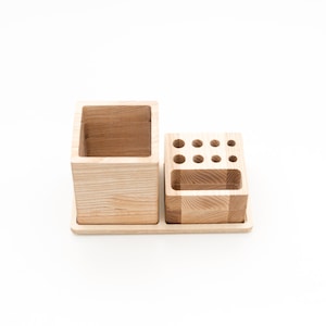 May include: A wooden desk organizer with two sections. One section is a large square cup, and the other has multiple small holes for pens and pencils. The organizer is made of light-colored wood and sits on a rectangular base.