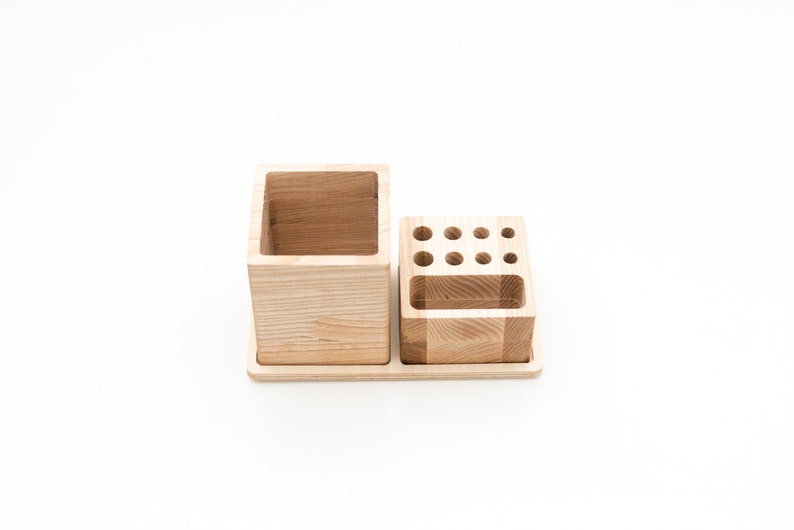 May include: A wooden desk organizer with two sections. One section is a large square cup, and the other has multiple small holes for pens and pencils. The organizer is made of light-colored wood and sits on a rectangular base.