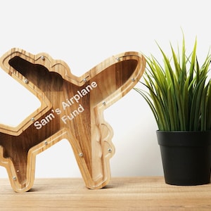 May include: Wooden airplane shaped piggy bank with a slot on top and a clear plastic window on the side. The piggy bank is painted brown and has the words "Sam's Airplane Fund" engraved on the side.