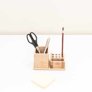 May include: A wooden desk organizer with two compartments. The left compartment holds a pair of black scissors and two silver pens. The right compartment has a single brown pencil standing upright. A yellow sticky note is on the white surface.