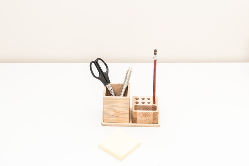 May include: A wooden desk organizer with two compartments. The left compartment holds a pair of black scissors and two silver pens. The right compartment has a single brown pencil standing upright. A yellow sticky note is on the white surface.