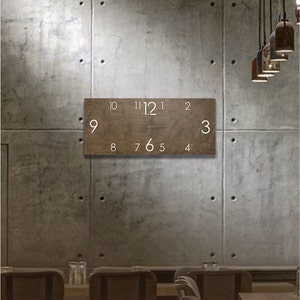 K&ouml;nnte beinhalten: Eine braune Holzuhr mit wei&szlig;en Zahlen an einer Betonwand. Die Uhr h&auml;ngt an der Wand &uuml;ber einem Tisch mit St&uuml;hlen.