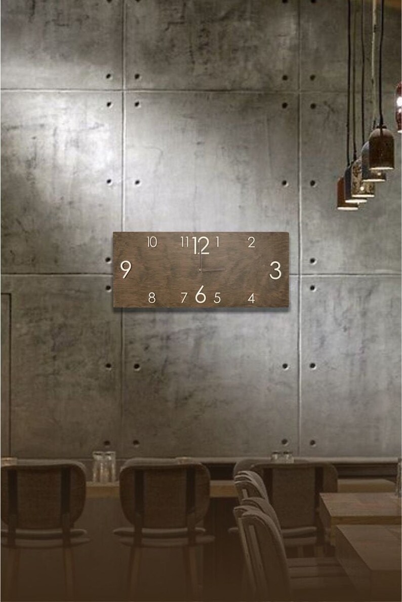 K&ouml;nnte beinhalten: Eine braune Holzuhr mit wei&szlig;en Zahlen an einer Betonwand. Die Uhr h&auml;ngt an der Wand &uuml;ber einem Tisch mit St&uuml;hlen.