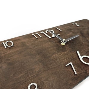 K&ouml;nnte beinhalten: Eine Holzuhr mit silbernen Metallziffern und Zeigern. Das Zifferblatt der Uhr ist dunkelbraun mit einer Holzmaserung. Die Ziffern sind erhaben und die Zeiger sind d&uuml;nn und elegant. Die Uhr ist auf 12:00 Uhr eingestellt.