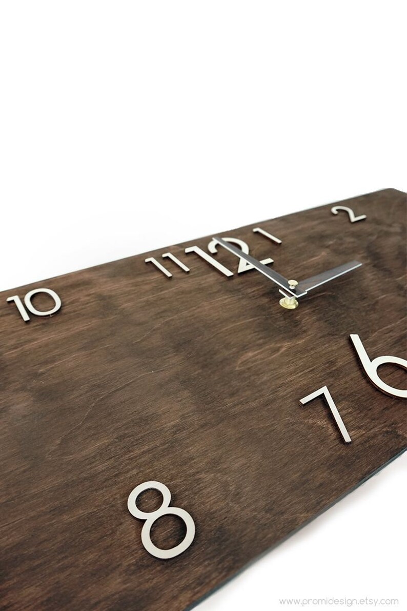 K&ouml;nnte beinhalten: Eine Holzuhr mit silbernen Metallziffern und Zeigern. Das Zifferblatt der Uhr ist dunkelbraun mit einer Holzmaserung. Die Ziffern sind erhaben und die Zeiger sind d&uuml;nn und elegant. Die Uhr ist auf 12:00 Uhr eingestellt.